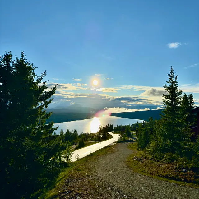 En svingete grusvei som leder ned mot en innsjø, med solen som speiler seg i vannet under en blå himmel.