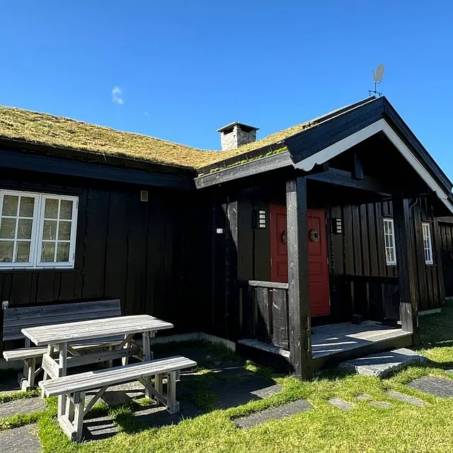En svart trehytte med rød inngangsdør og et uteområde med benker og bord, under en klar blå himmel.