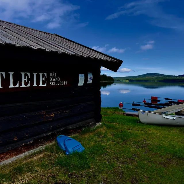 En utleiehytte ved en innsjø med kanoer på en brygge og klare refleksjoner av himmel og åser i vannet.