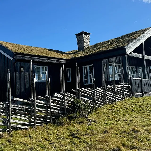 En svart trehytte med gress på taket og en steinskorstein, omgitt av et skrått gressdekket terreng og et gjerde laget av trestolper.
