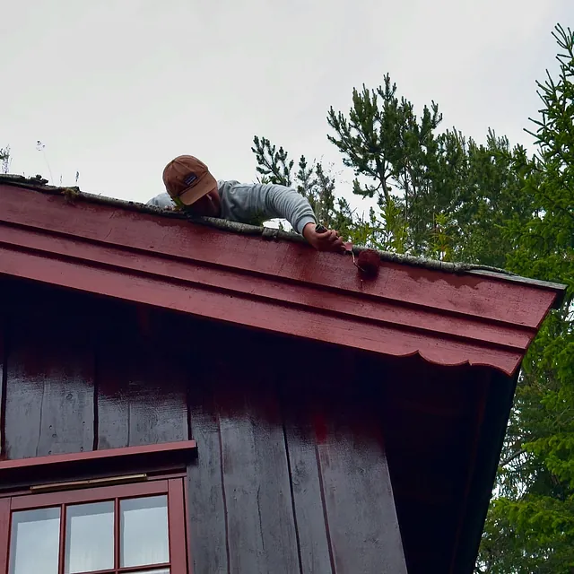 En mann maler kanten av et hyttetak rødt, med et tre i bakgrunnen.